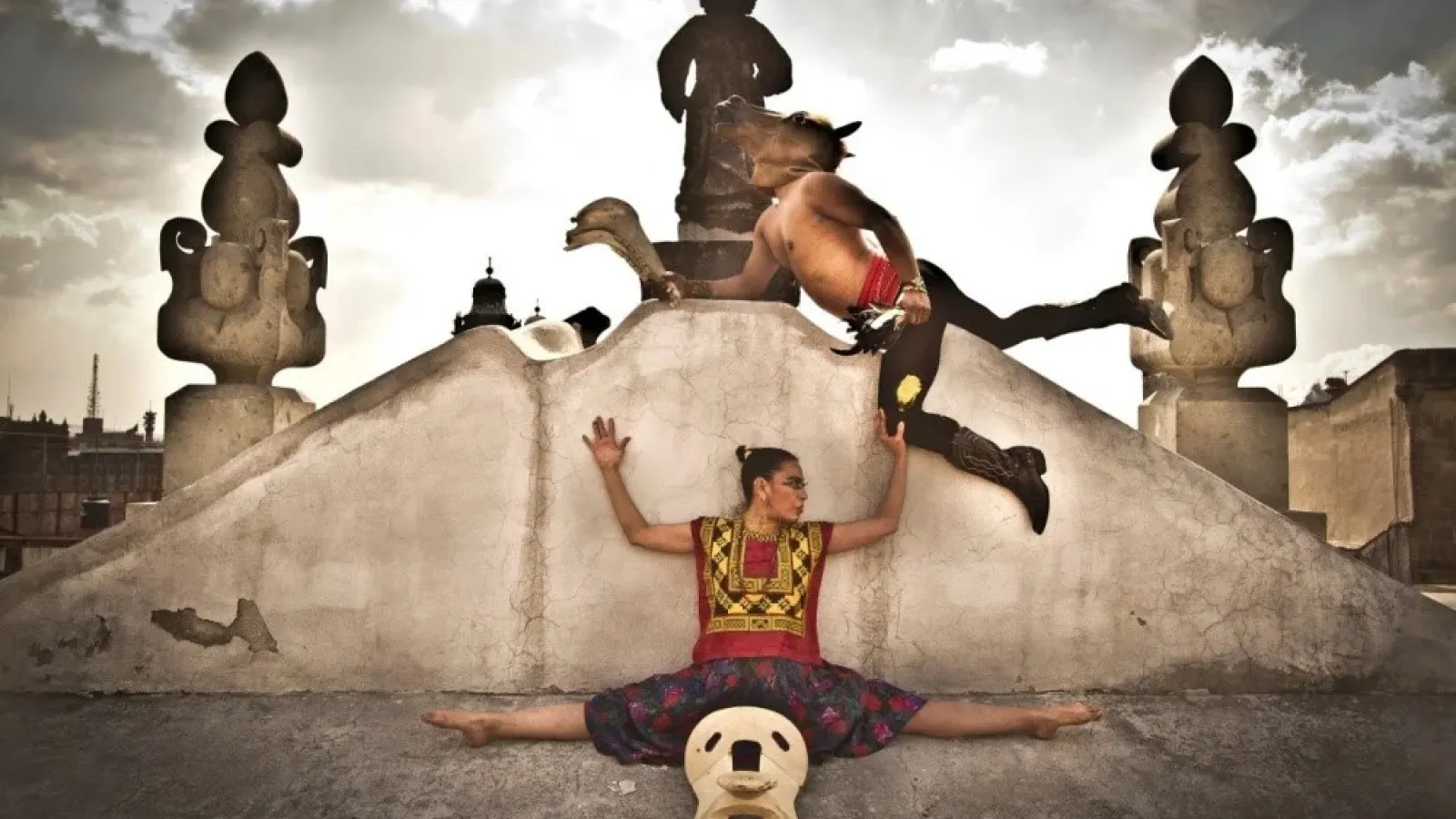 A dancer doing the splits on the ground in front of a monument, with another dancer leaping over hear head wearing a horse mask. 