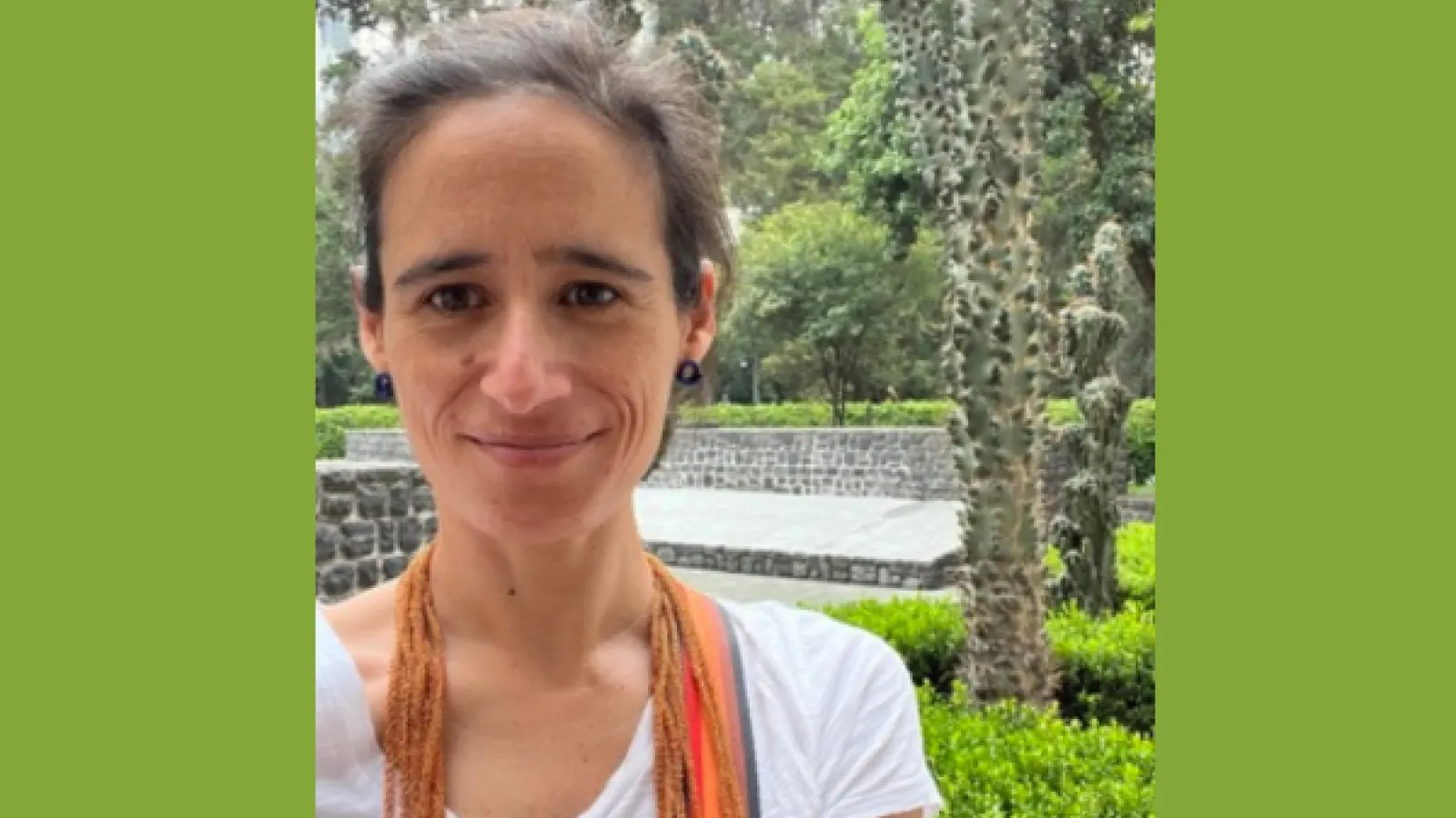 Headshot of Ines Valdez. She has brown hair that is pulled back, is wearing a white vneck tshirt and an orange scarf. 