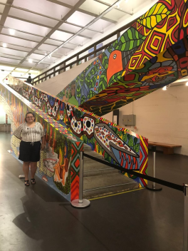 a photo of Mello at MASP standing beside the work made by Indigenous artist collective MAHKU at the Museu de Arte de Sao Paulo