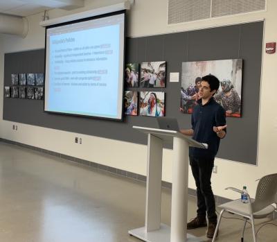 Kevin stands at a podium at the front of the room with a screen behind him. There are large photographs on the wall, but the subjects are blurry.