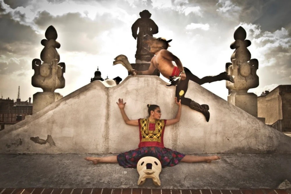 A dancer doing the splits on the ground in front of a monument, with another dancer leaping over hear head wearing a horse mask.