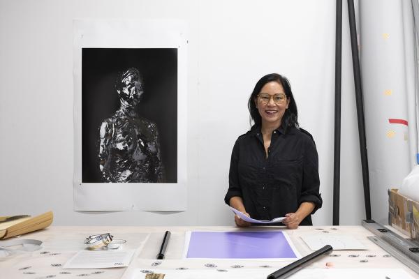 Gina Osterloh looks at the camera with her work table and tools in front of her. One of her photographs is hung on the wall to her right. 