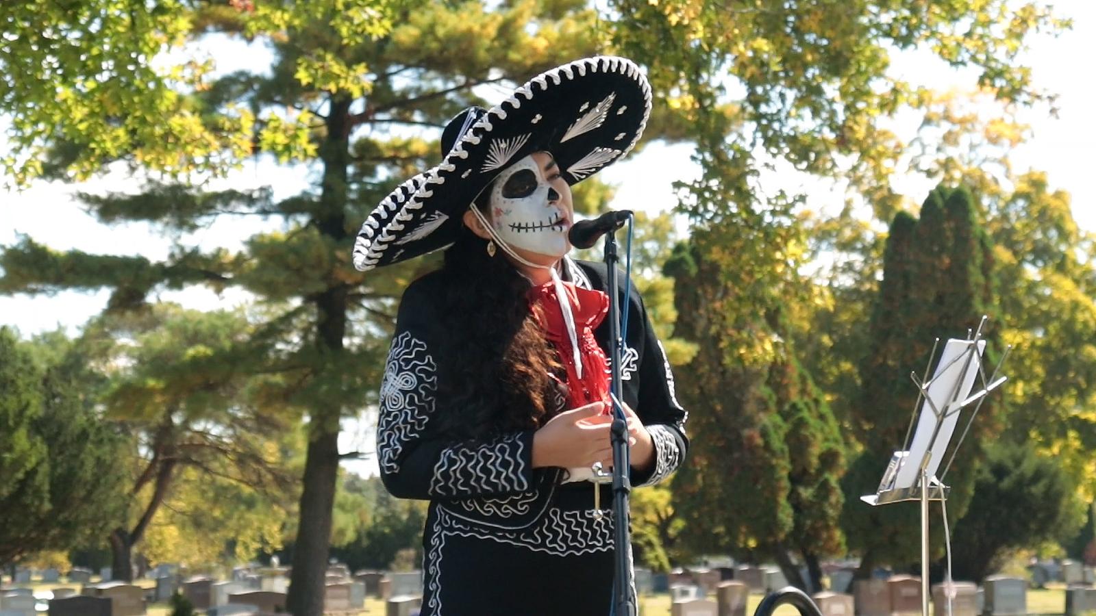 A woman in a mariachi uniform and face makeup sings into a microphone. 
